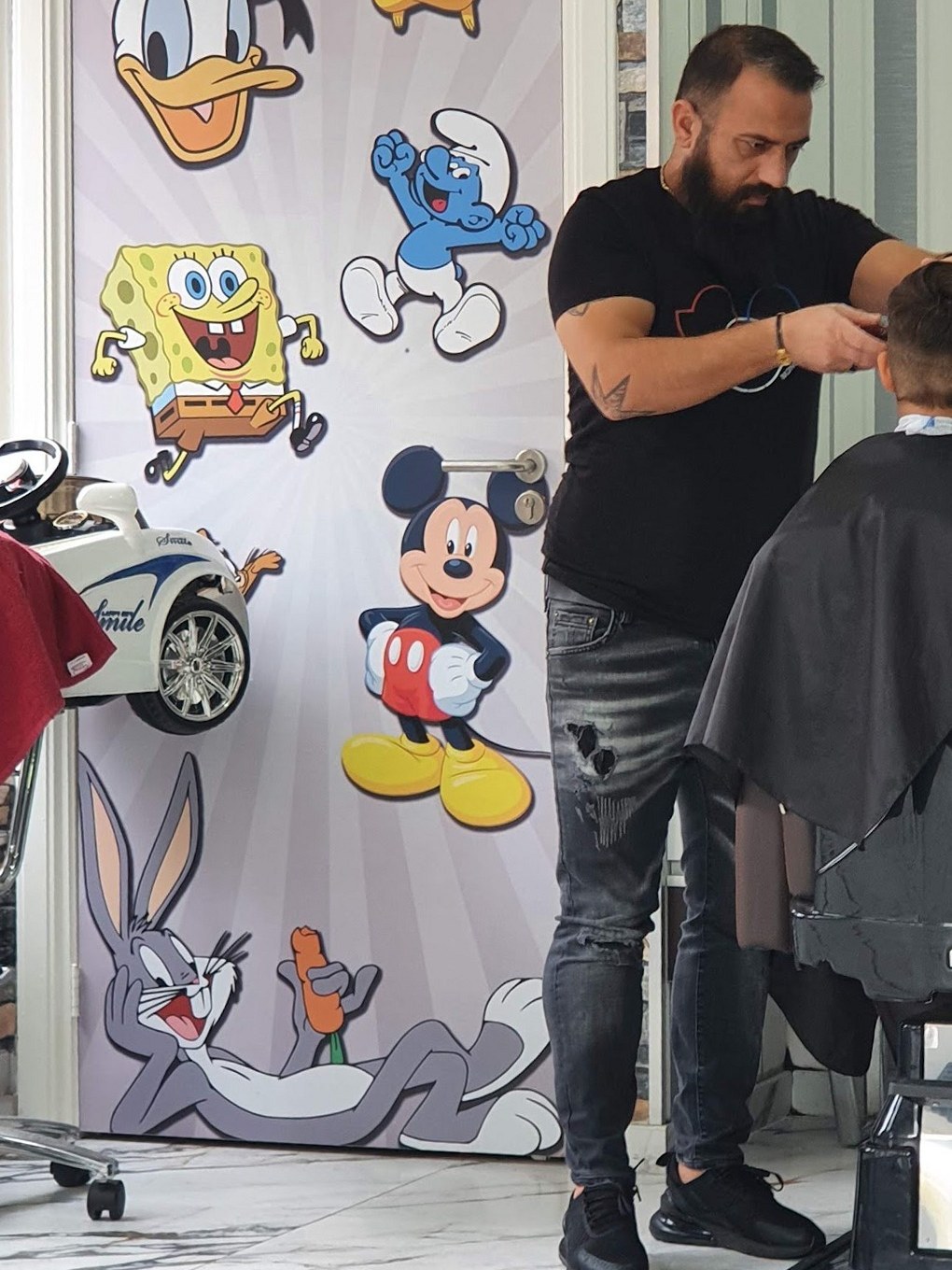 Een kapper aan het werk bij een jongen in de stoel, naast een deur vol tekenfilmfiguren